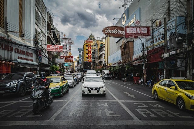 Une rue animée en Thaïlande avec un mélange de voitures, taxis et motos sous un ciel nuageux, mettant en valeur l'effervescence de la circulation urbaine.