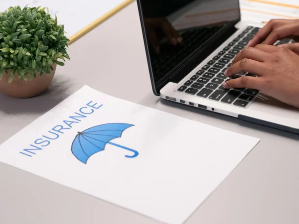 Personne tapant sur un ordinateur portable à côté d'un document intitulé 'Assurance' avec une icône de parapluie bleu, symbolisant la couverture et la protection.