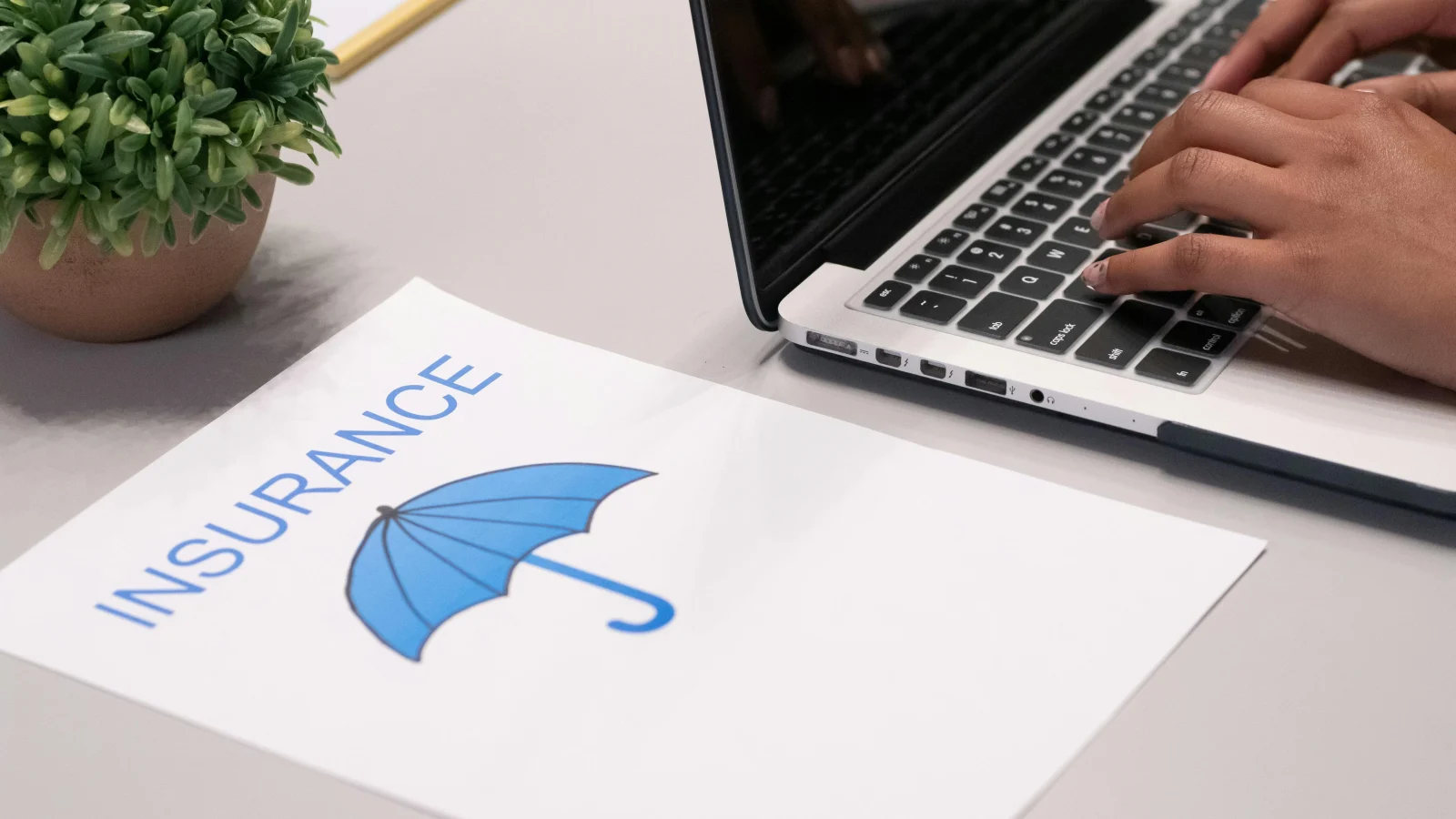 A person typing on a laptop next to a document labeled 'Insurance' with a blue umbrella icon, representing protection and coverage.