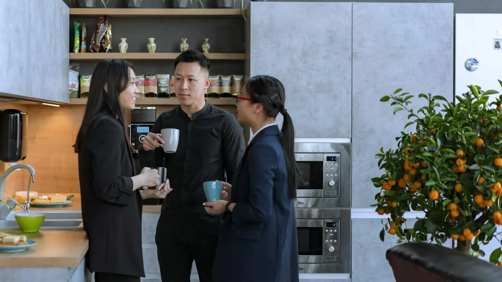 Trois collègues discutant autour d'une tasse de café dans une cuisine moderne d'un bureau, avec des étagères décorées de plantes et des appareils électroménagers intégrés. Une ambiance détendue, propice à l'échange informel entre professionnels.