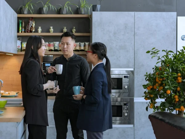 Trois collègues discutant autour d'une tasse de café dans une cuisine moderne d'un bureau, avec des étagères décorées de plantes et des appareils électroménagers intégrés. Une ambiance détendue, propice à l'échange informel entre professionnels.