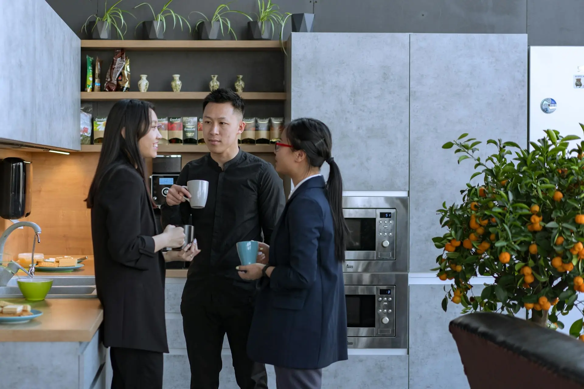 Trois collègues discutant autour d'une tasse de café dans une cuisine moderne d'un bureau, avec des étagères décorées de plantes et des appareils électroménagers intégrés. Une ambiance détendue, propice à l'échange informel entre professionnels.