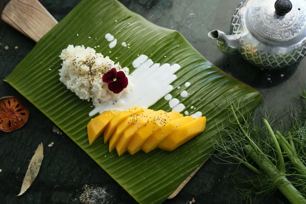 A traditional Thai dessert with sweet sticky rice, fresh mango slices, and coconut milk, elegantly served on a banana leaf with sesame seeds and a flower garnish.