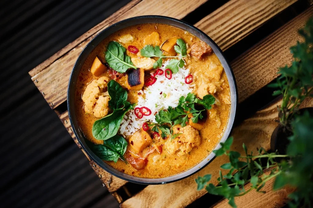 A bowl of creamy Massaman curry with tender meat, potatoes, vegetables, and white rice, garnished with fresh cilantro, red chili slices, and greens.