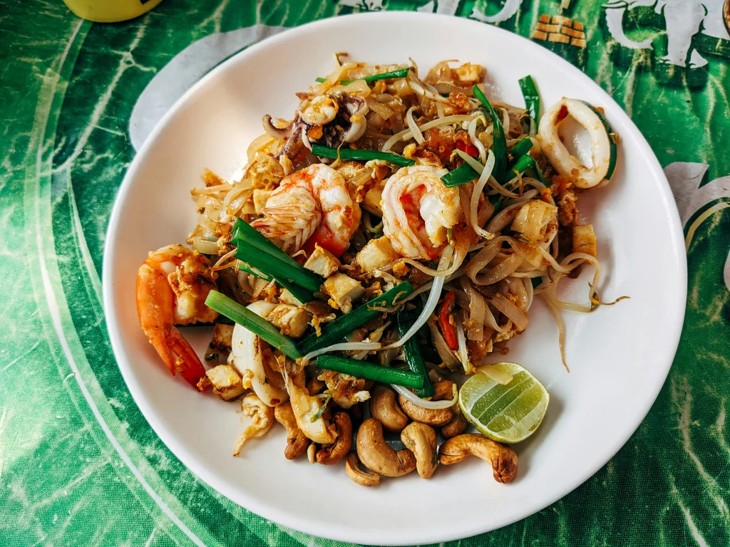 A plate of stir-fried rice noodles with shrimp, tofu, bean sprouts, green onions, and a lime wedge, served on a vibrant tablecloth with cashews on the side.