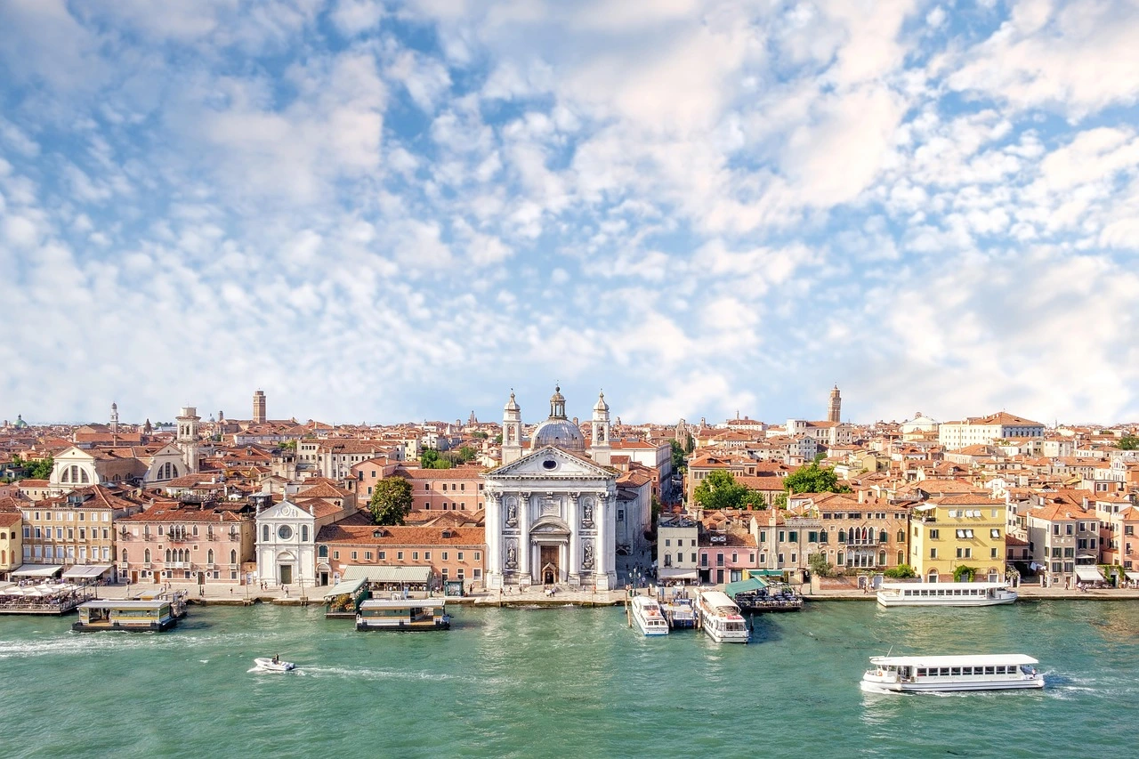 A stunning view of Venice’s waterfront, featuring historic architecture, domed churches, and boats navigating the canal under a bright blue sky.
