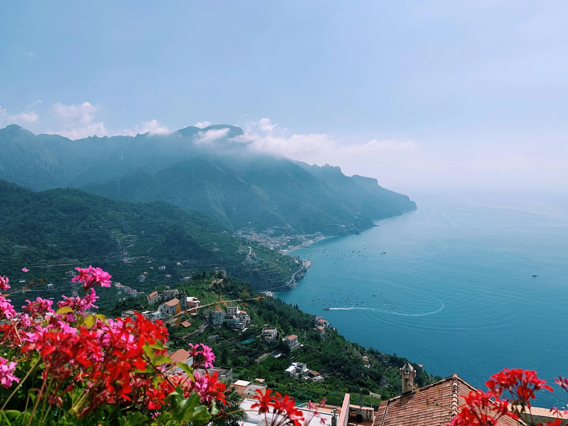 Scenic view of the Amalfi Coast with colourful flowers in the foreground, coastal villages below, and misty mountains rising along the Mediterranean Sea.
