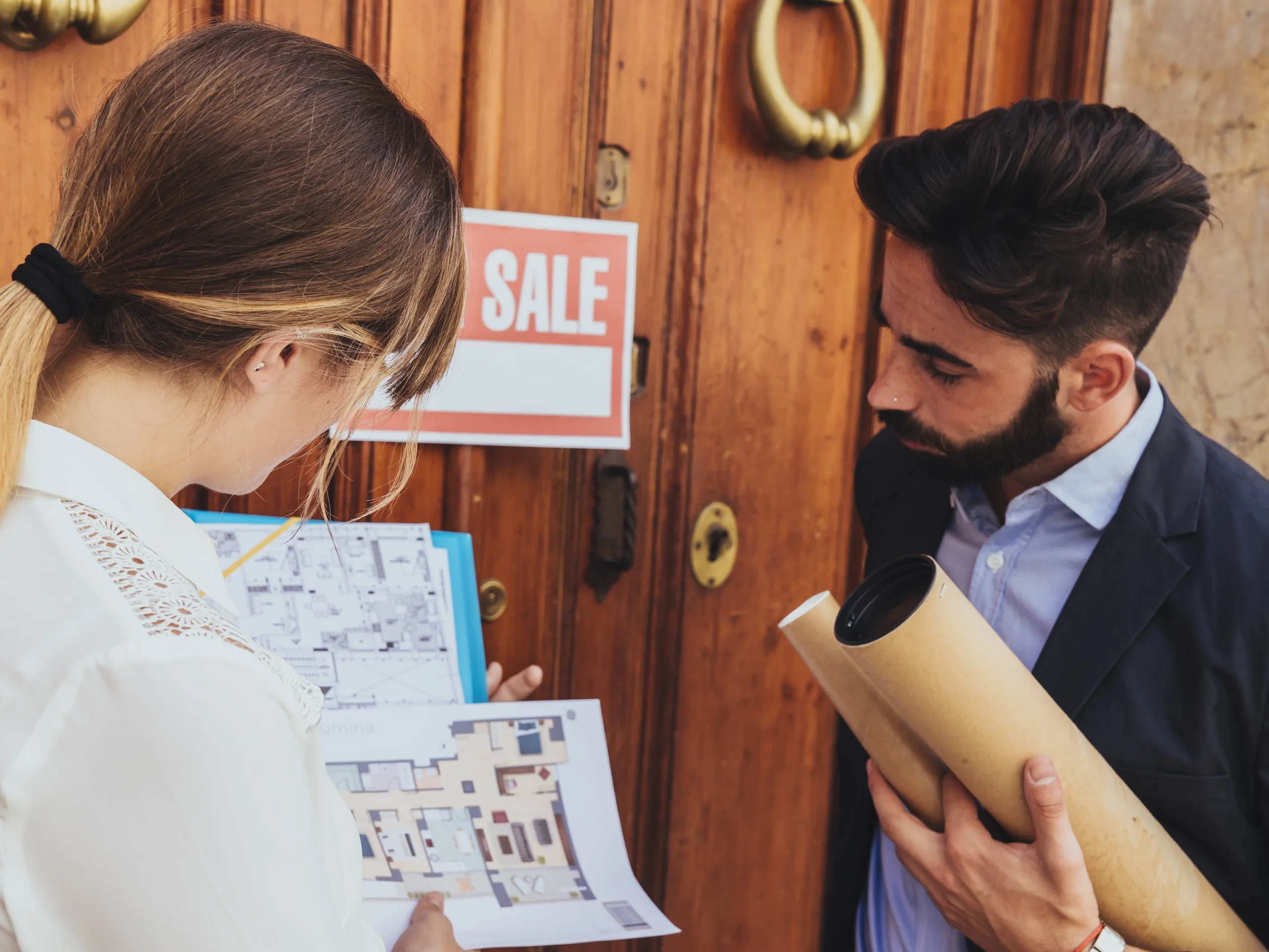 Agent immobilier aidant des clients avec des plans et des visites, devant une maison avec un panneau à vendre pour acheter une maison en Espagne.