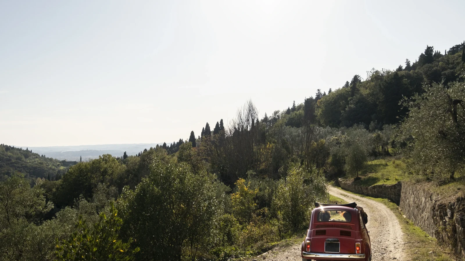Vintage red car driving through the countryside during a scenic trip to Italy, surrounded by olive trees and Tuscan hills.