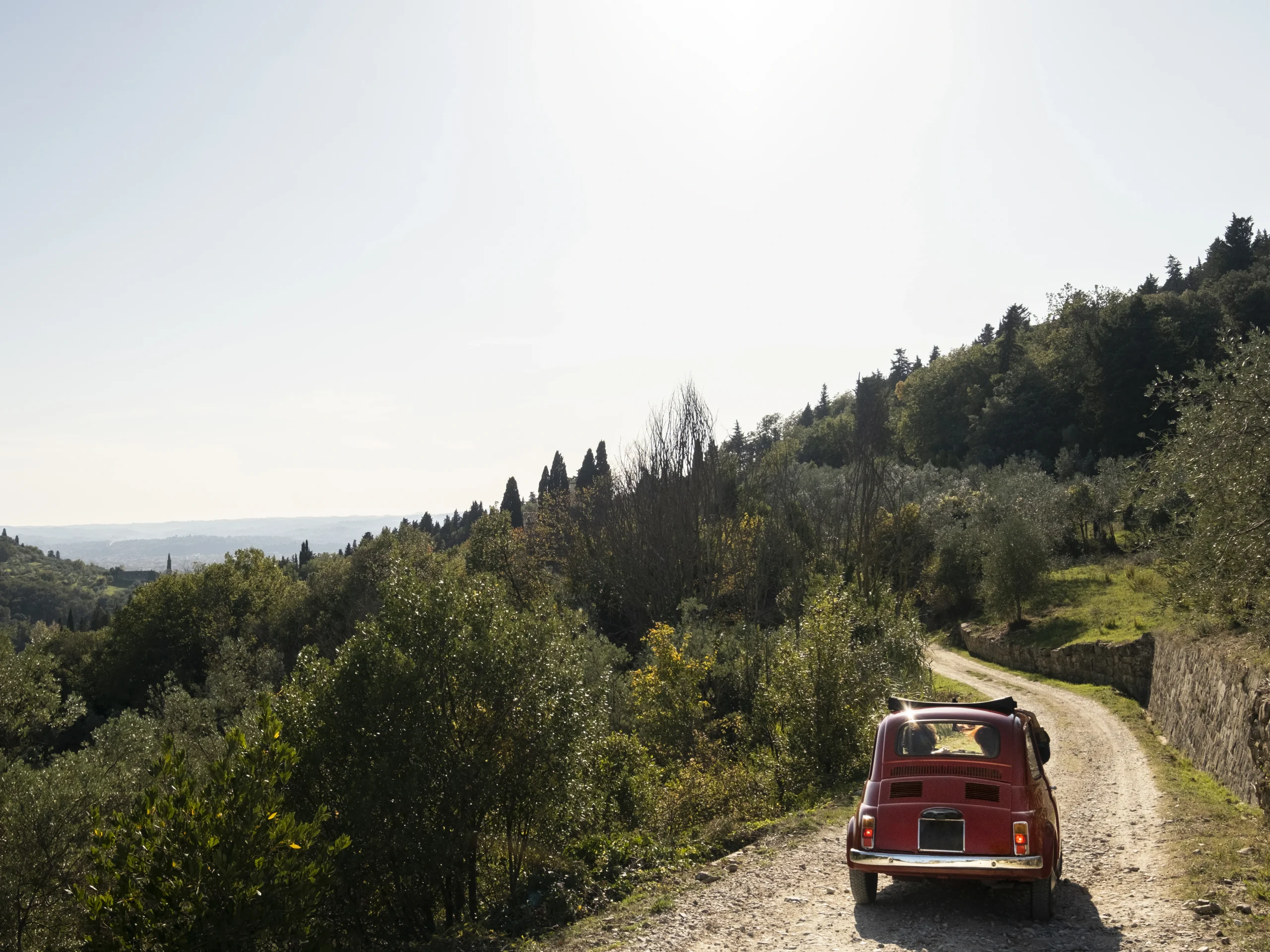 Vintage red car driving through the countryside during a scenic trip to Italy, surrounded by olive trees and Tuscan hills.