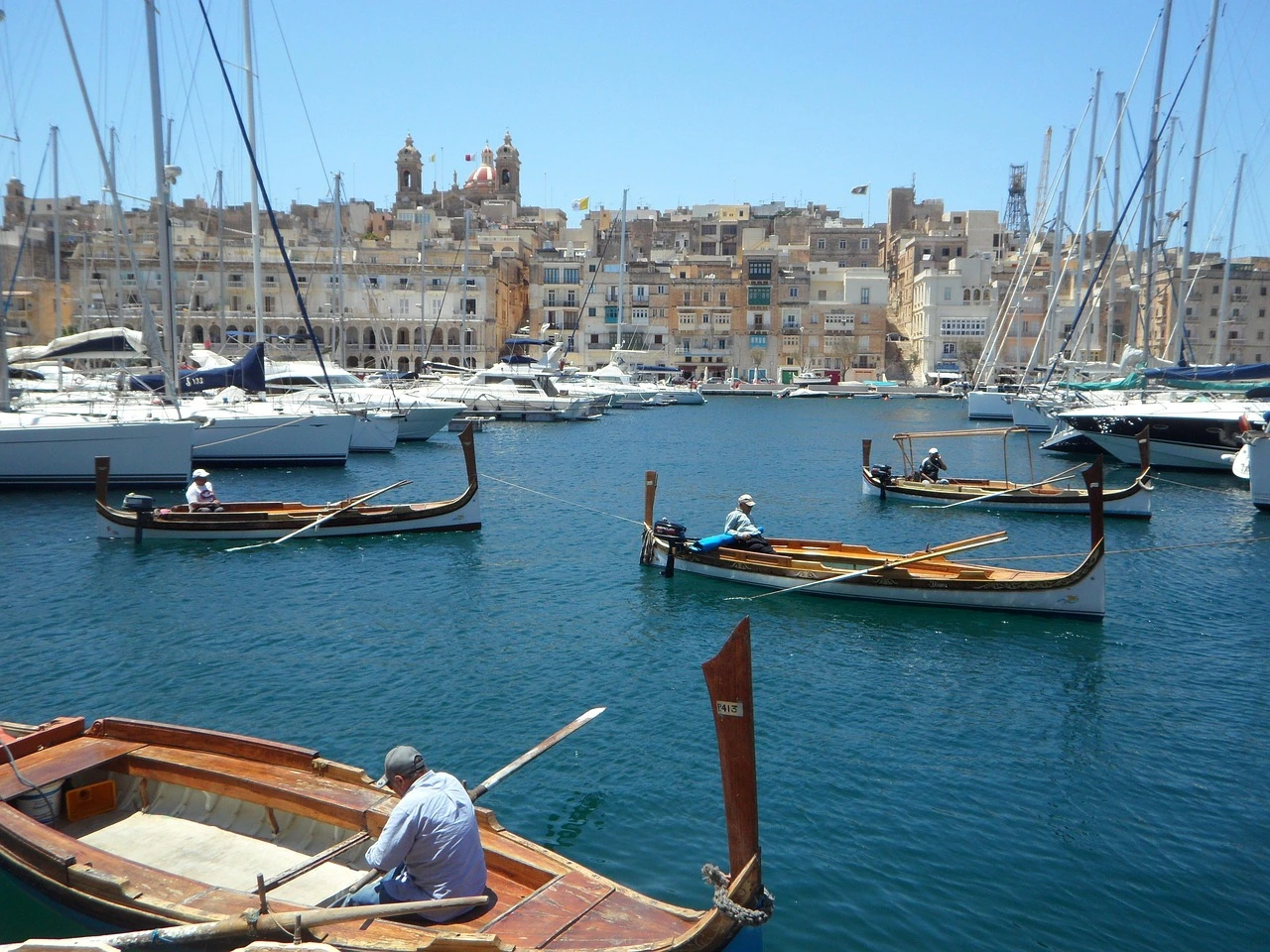 Traditional Maltese boats and yachts docked in a peaceful harbour with historic buildings in the background, showcasing daily life in Valletta.