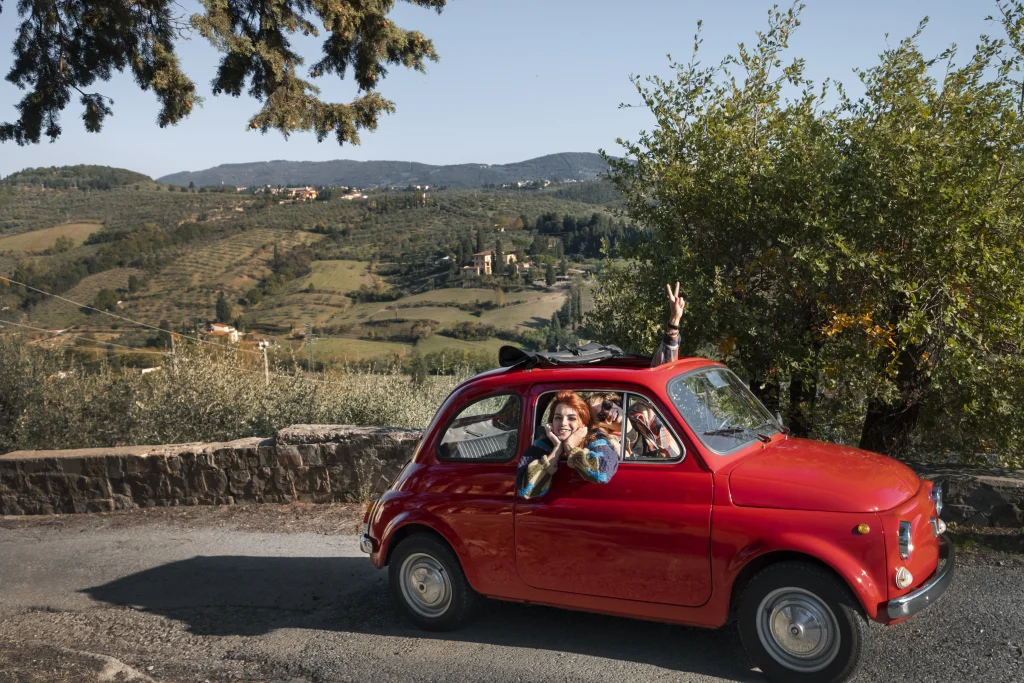 Joyful travellers in a red Fiat on a road trip to Italy, exploring charming villages and green landscapes in Tuscany.