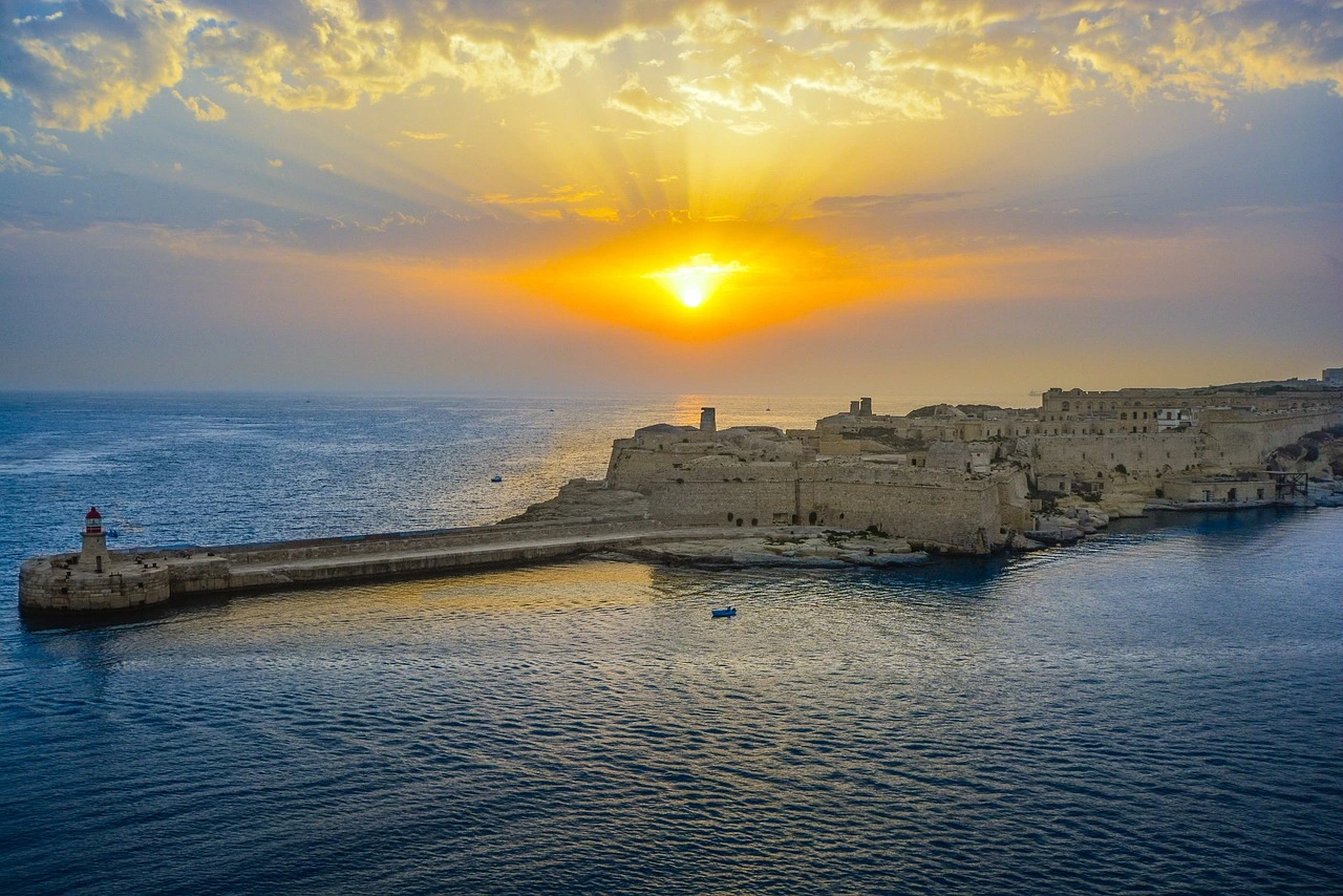 Lever de soleil doré sur la mer et les anciennes fortifications à Malte, projetant une lumière dramatique sur l'eau et les structures en pierre anciennes.