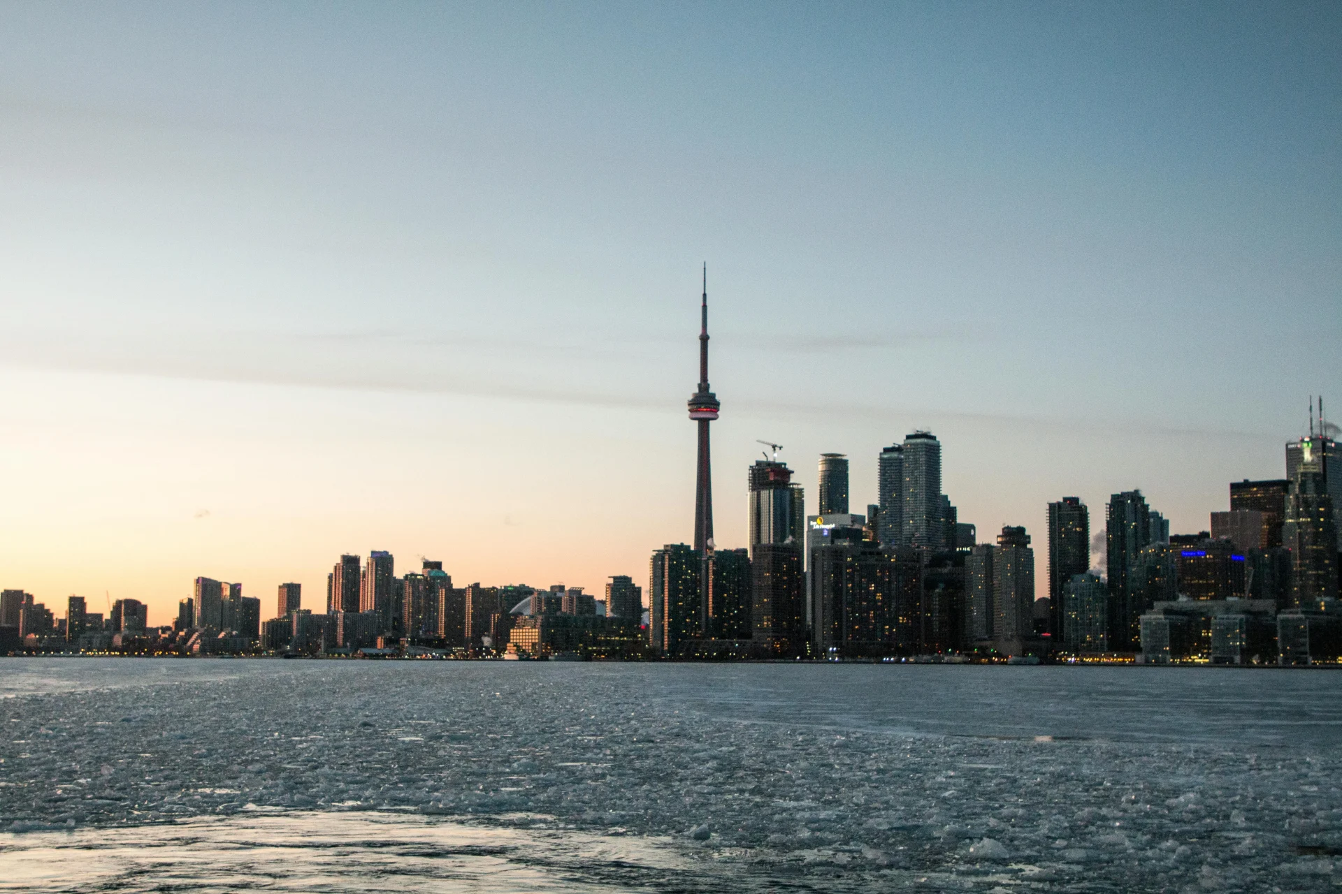 Skyline de Toronto au coucher du soleil avec la tour CN bien visible, les lumières de la ville qui s’allument et les eaux glacées du lac au premier plan.