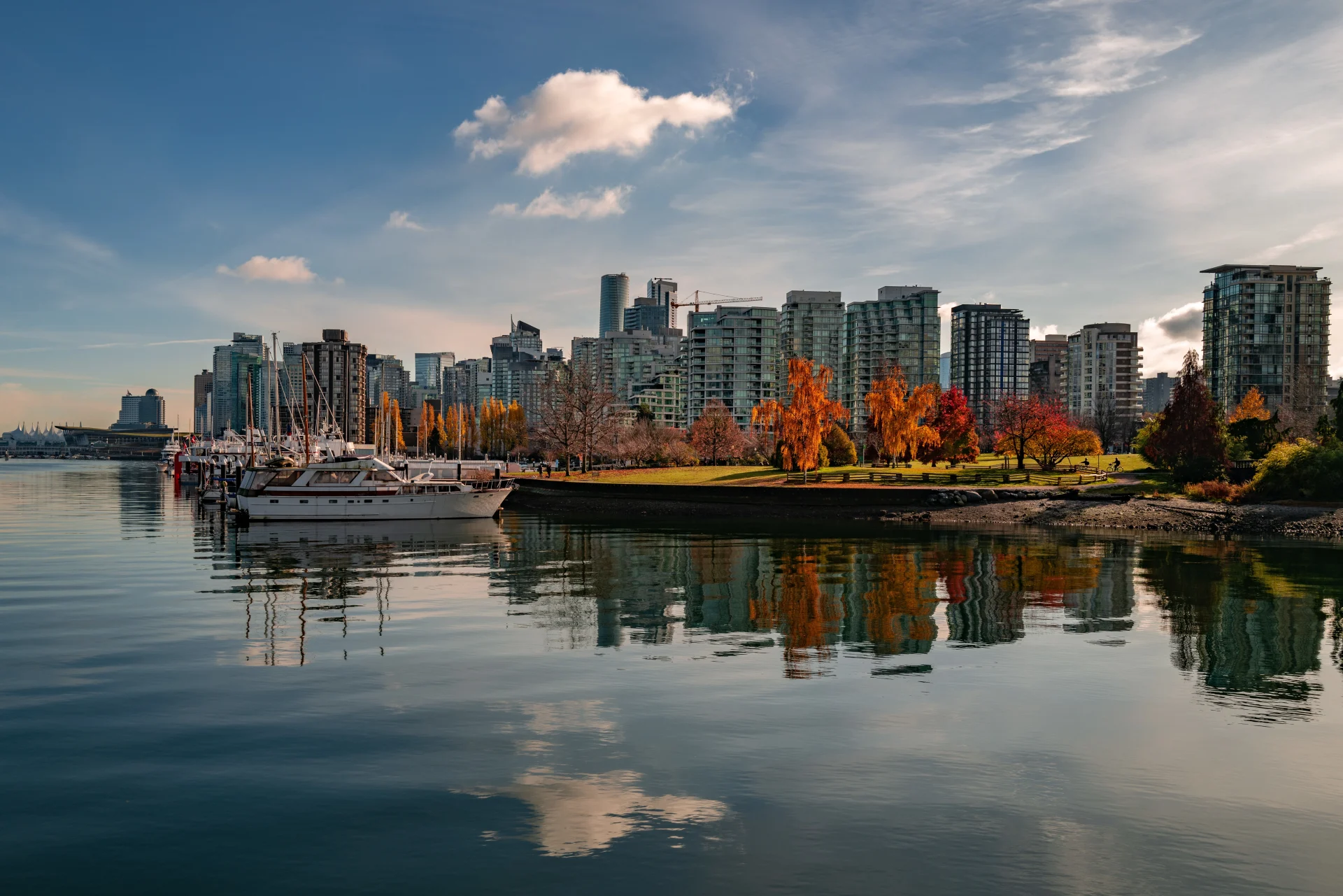 Vue panoramique de la skyline de Vancouver avec des yachts, des arbres d’automne et des reflets sur un port calme sous un ciel bleu éclatant.