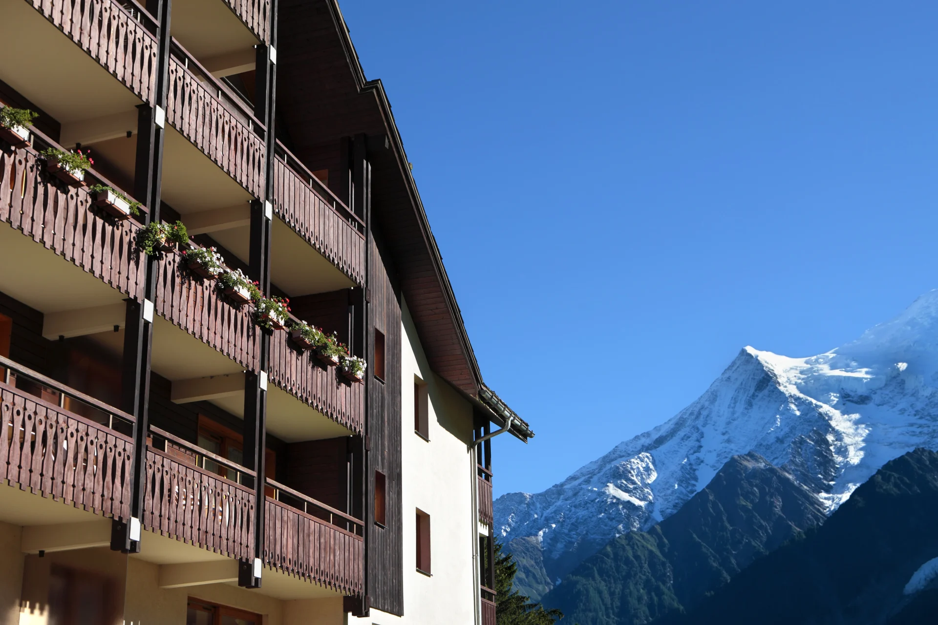 Traditional wooden Swiss apartment balconies with flower boxes, set against a backdrop of snowy Alpine peaks and a clear blue sky.