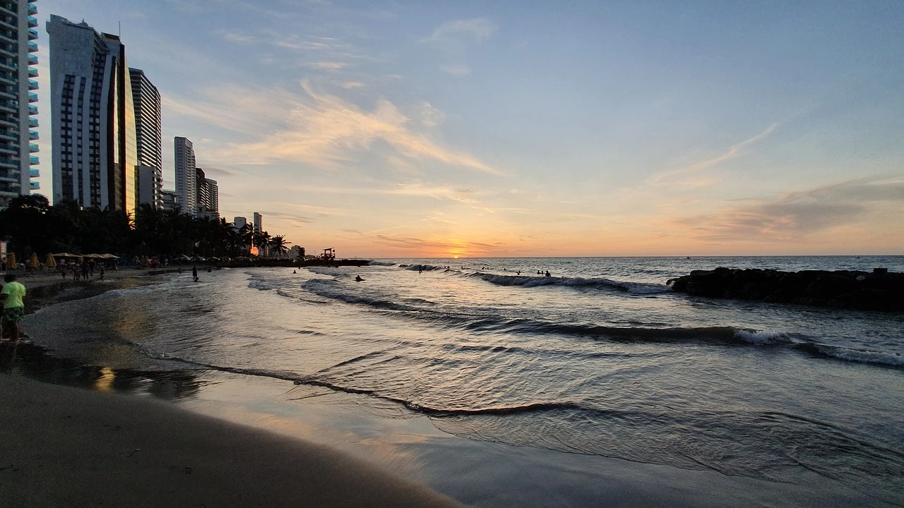 Les vagues roulent tandis que le soleil se couche derrière des tours modernes en bord de mer, peignant le ciel de teintes dorées et projetant des reflets sur le sable.