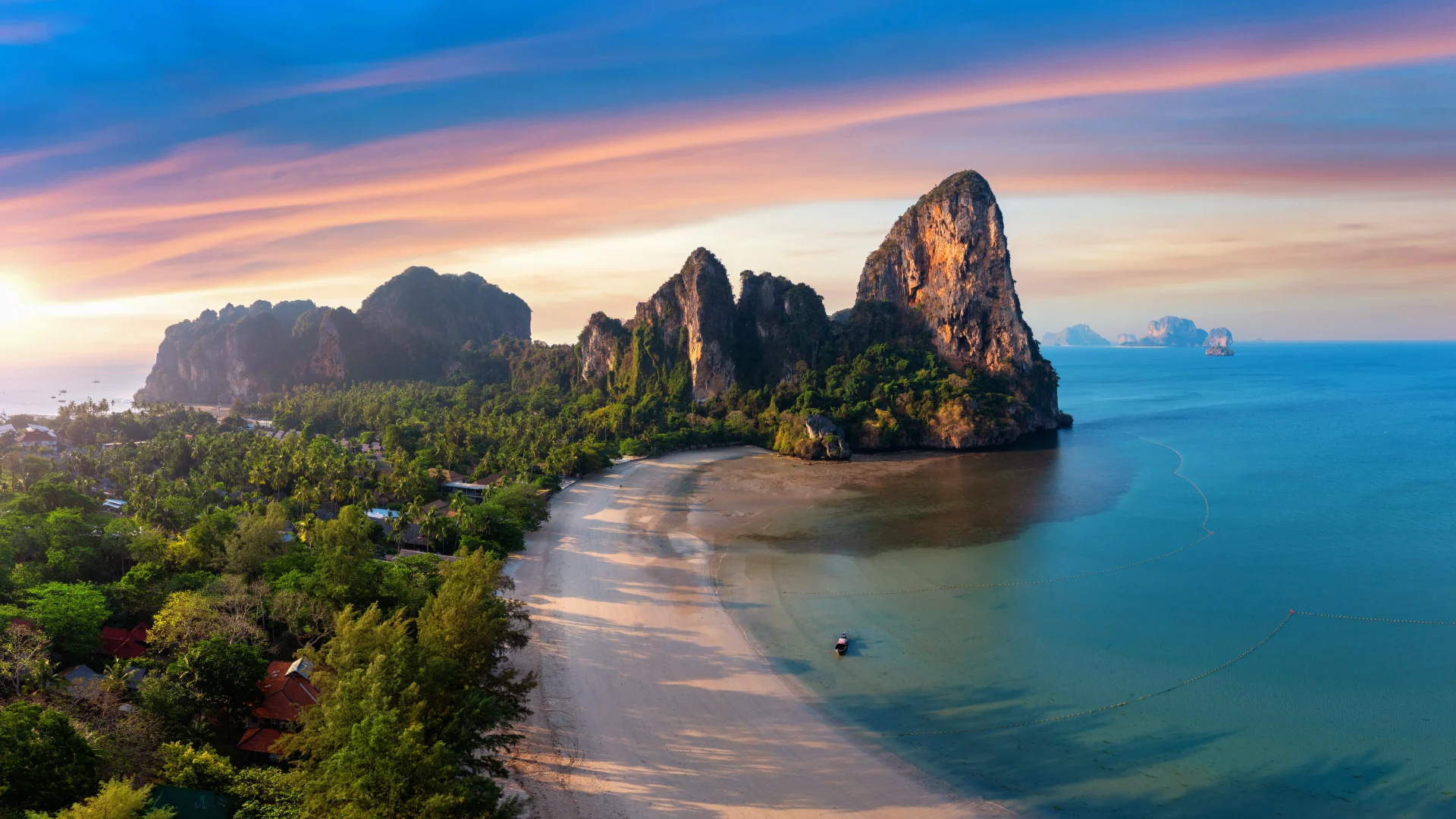 Scenic view of Railay Beach in Krabi, Thailand, with limestone cliffs, turquoise sea, lush forest, and a colorful sunset sky over the coastline.