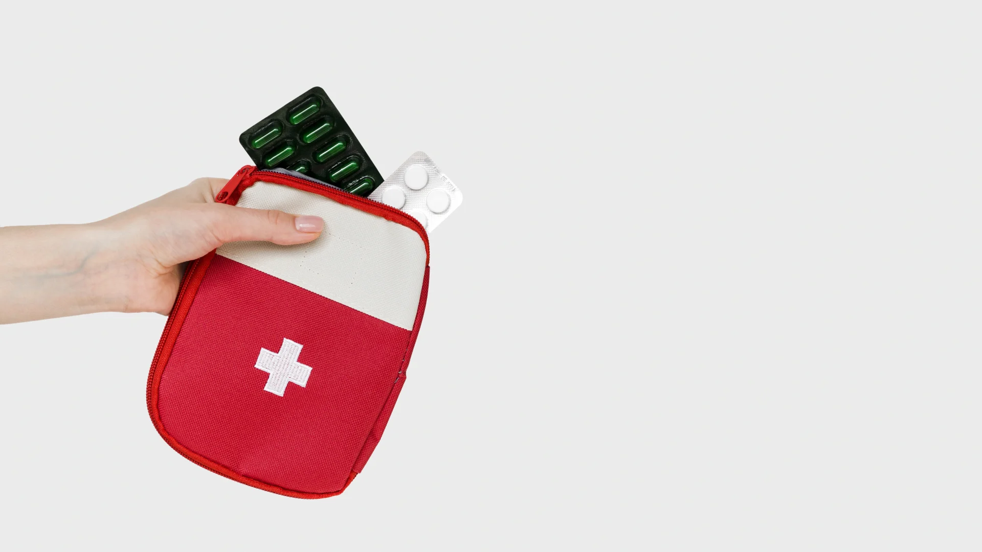 Main tenant une trousse de secours rouge avec une croix blanche, contenant des plaquettes de médicaments, sur un fond gris clair.