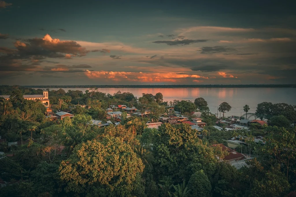 Vue pittoresque d’un village colombien paisible, entouré d’une jungle luxuriante et d’une rivière calme au coucher du soleil, sous des nuages teintés d’orange.