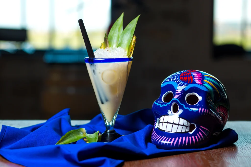 A creamy cocktail garnished with pineapple leaves and ice in a blue-rimmed glass sits beside a colorful, hand-painted blue Mexican skull on a deep blue cloth.