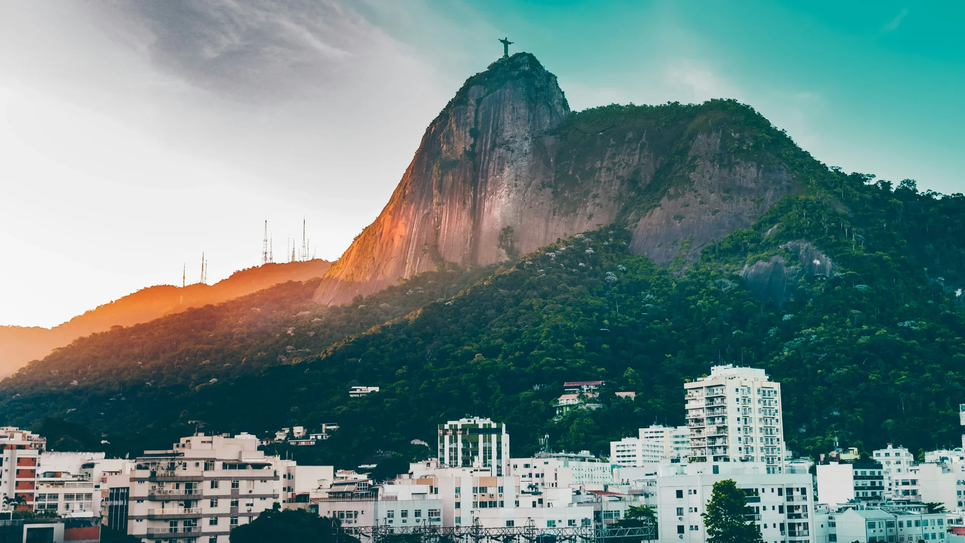 View of Corcovado Mountain with Christ the Redeemer statue overlooking residential buildings in Rio de Janeiro at sunset.