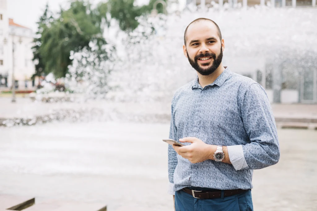 Smiling expat in Portugal checking tax info on his phone near a city fountain