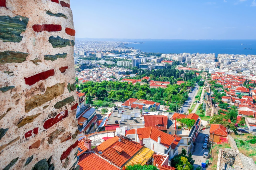 Vue panoramique de Thessalonique depuis les anciennes murailles, avec toits rouges, végétation dense et mer Égée en toile de fond. Une des meilleures villes en Grèce pour allier culture et vie côtière.
