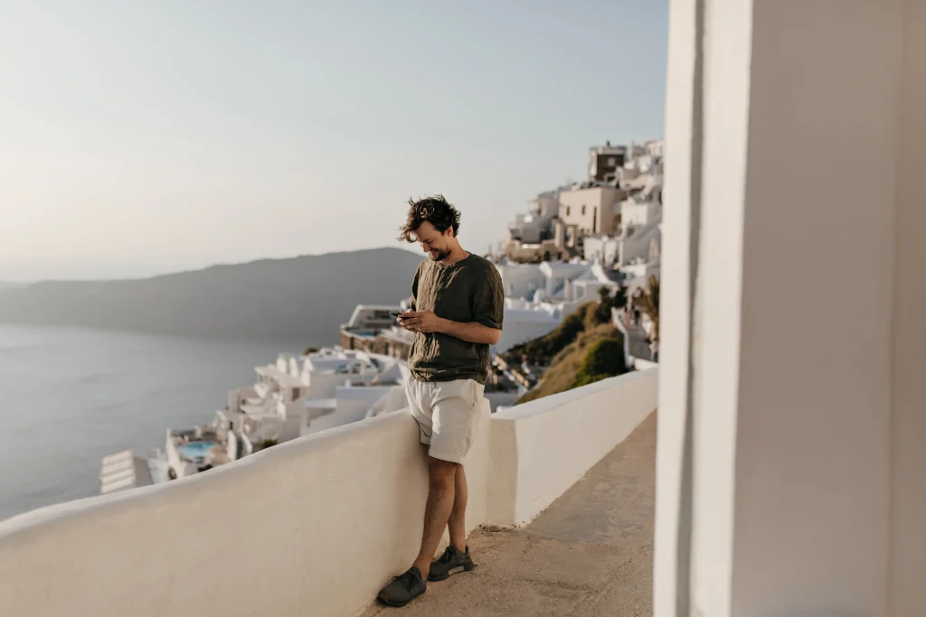 Jeune homme en short et t-shirt profitant d’une vue spectaculaire sur les falaises de Santorin et la mer, entouré des célèbres maisons blanches perchées à flanc de colline. Un moment paisible au cœur d’un décor cycladique iconique.