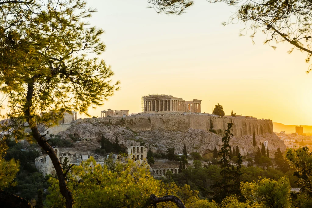 Vue panoramique sur Athènes depuis les hauteurs, avec l’Acropole en toile de fond, des bâtiments blancs à perte de vue et un ciel clair d’été — parfait pour illustrer la vie urbaine en Grèce.
