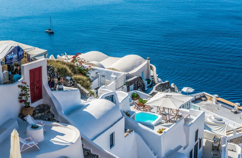  Vue emblématique des toits blancs et terrasses ensoleillées de Santorin, surplombant la mer Égée d’un bleu profond. Une scène typique des Cyclades avec ses ruelles sinueuses, ses fleurs grimpantes et une ambiance méditerranéenne sereine.