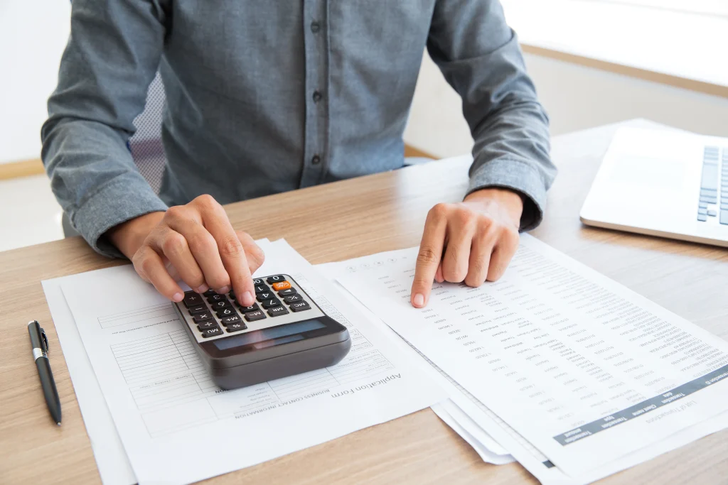 A man in a dress shirt calculates the cost of living in Turkey using a calculator and financial documents on a desk.