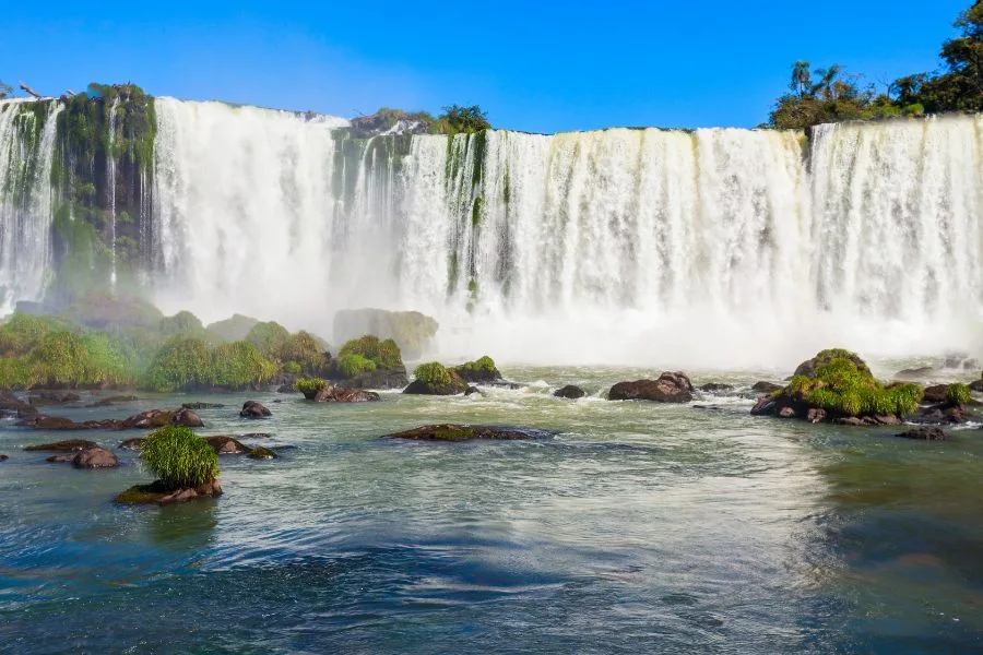 The impressive Iguaçu Falls on the Brazilian side, with powerful cascades and mist rising above the water under a blue sky.